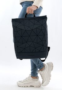 Svart ryggsäck med geometriskt mönster och texturerad yta, topphandtag och justerbara axelremmar. Märkesetikett synlig längst ner.