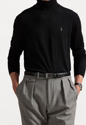 Homme portant un pull à col roulé noir avec un petit logo, un pantalon gris plissé, une ceinture en cuir noire et une montre-bracelet, les mains dans les poches.