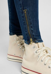 Jean skinny bleu en denim avec un détail de zip doré aux chevilles, associés à des baskets montantes en toile beige avec des accents rouges et navy.