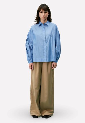 Femme debout, face à l'avant, portant une chemise ample à rayures bleues et un pantalon beige à jambes larges, sur un fond uni.