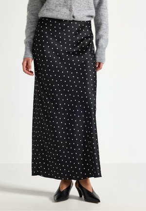 Sort satin maxi nederdel med hvide polkaprikker båret med grå cardigan og sorte hæle på en stående person.
