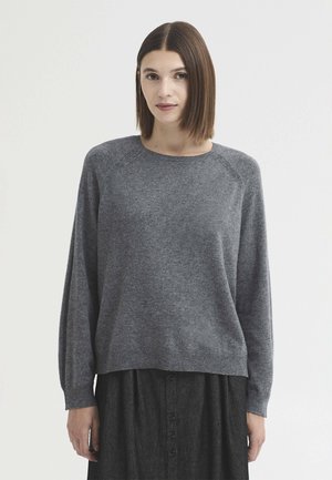 Femme aux cheveux bruns lisses portant un pull gris à manches longues et une jupe sombre boutonnée, debout devant un fond blanc uni.