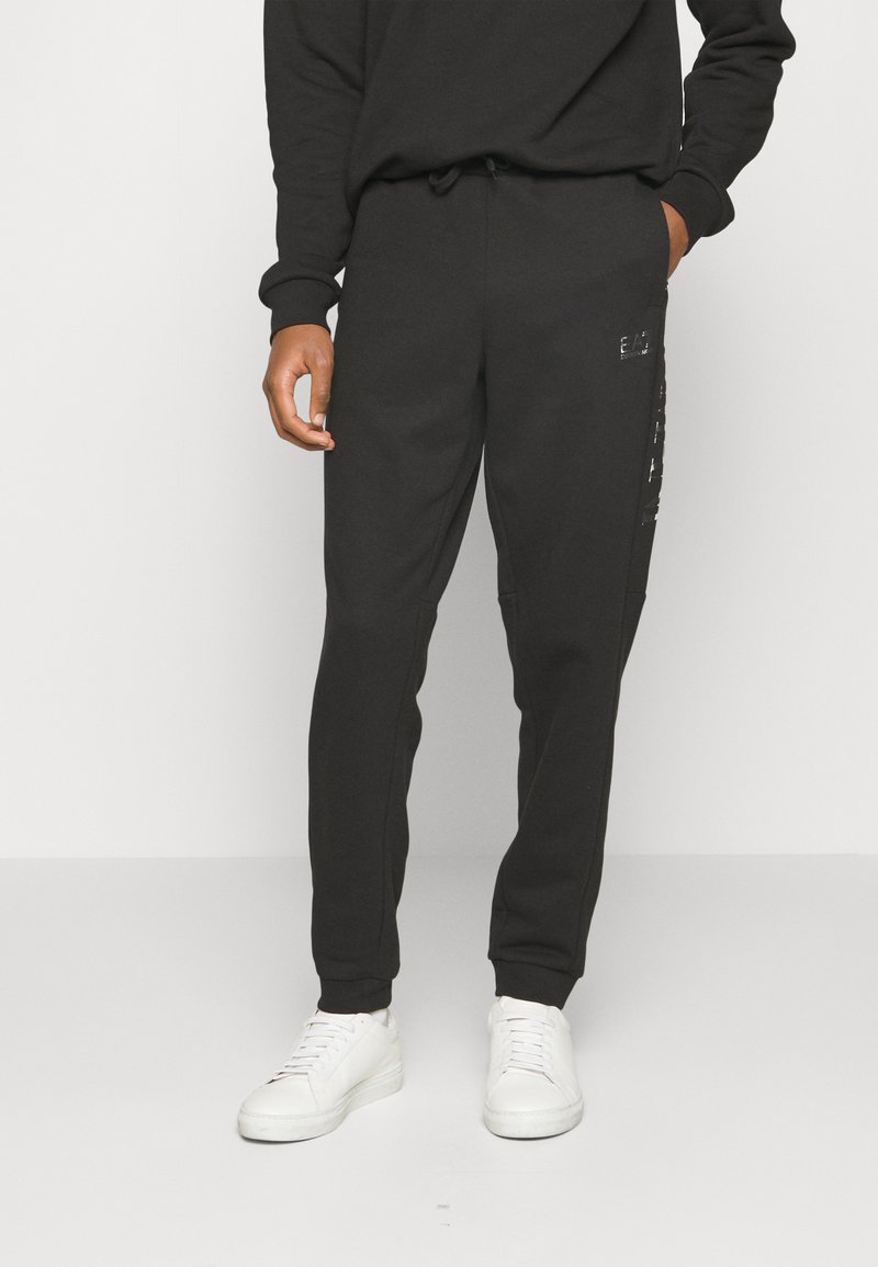 Pantalones de chándal negros de corte ajustado, con cintura elástica y bolsillos laterales. Incluye un detalle de logo brillante a lo largo de la pierna izquierda. Tejido de mezcla de algodón.