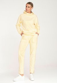 Żółta bluza z kapturem i pasujące spodnie z miękkiego materiału, z kieszenią kangurką oraz bocznymi rozporkami w spodniach, w połączeniu z białymi trampkami.