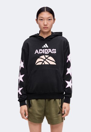 Ung person med glat mørkt hår bærer en sort Adidas-hættetrøje med lyserøde stjerner på ærmerne og olivengrønne Adidas-shorts.