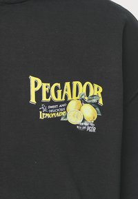 Felpa nera con una grafica colorata. Il testo include "PEGADOR" in giallo, con illustrazioni di limoni e la frase "Sweet and Delicious Lemonade".
