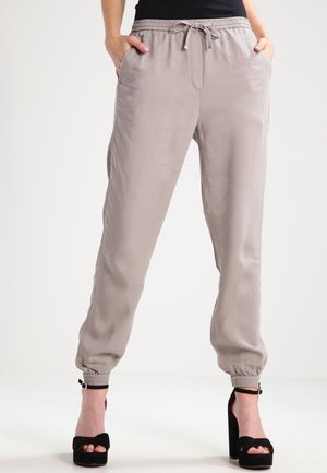 Frau trägt beigefarbene Jogginghosen mit Kordelzug und Seitentaschen, kombiniert mit schwarzen High-Heel-Sandalen vor weißem Hintergrund.