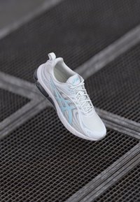 Hvid atletisk sko med lyseblå detaljer, mesh-overdel, polstret krave og gummisål, der har et strømlinet design og snøresystem.