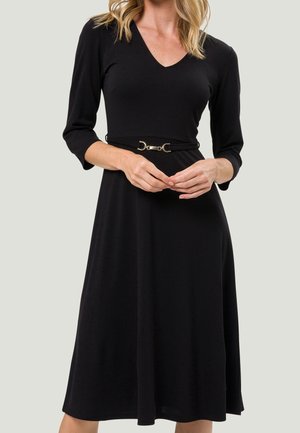 Femme portant une robe noire à encolure en V, manches trois-quarts et une ceinture fine avec une boucle dorée à la taille.