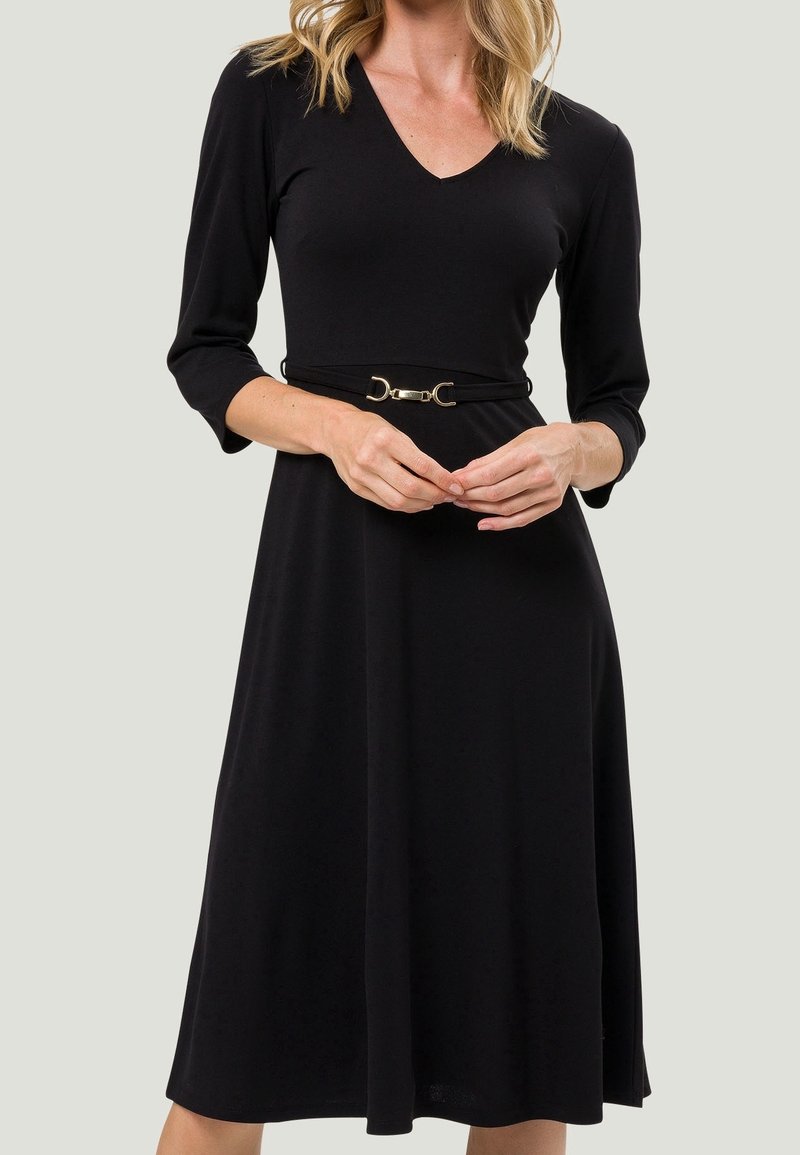 Femme portant une robe noire à encolure en V, manches trois-quarts et une ceinture fine avec une boucle dorée à la taille.