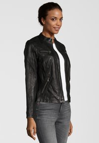 Schwarze Leder-Motorradjacke mit Stehkragen, Reißverschluss-Taschen und taillierter Passform. Getragen über einem weißen Hemd und kombiniert mit grauen Jeans.