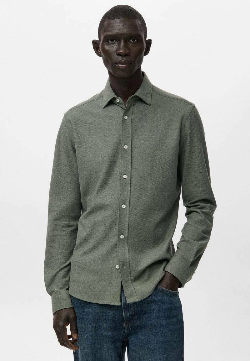 Camisa verde de manga larga con superficie texturizada, cierre de botones y cuello puntiagudo, combinada con jeans de mezclilla azul.