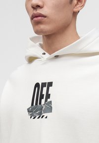 Jeune homme portant un sweat à capuche blanc avec un texte noir "OFF" partiellement caché par une bande de ruban gris au niveau de la poitrine.