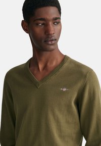 Olivgrön v-ringad tröja, stickad tyg, med en liten logotyp på bröstet, med en mjuk yta och ren finish.