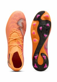 Kirkkaan oranssit jalkapallokengät, joissa on teksturoitu päällinen, edessä nauhat ja musta pohja, jossa on vaaleanpunaiset nastat. Mukana erottuvia suunnitteluelementtejä.