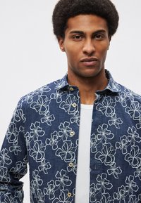 Τζιν πουκάμισο με λουλουδάτο μοτίβο σε ναυτικό μπλε με λευκή κεντητική. Διαθέτει μπροστινό μέρος με κουμπιά και άνετη εφαρμογή, φορεμένο πάνω από λευκή T-shirt.