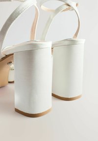 Valkoiset korkokengät, joissa on paksut paksut korot ja nilkkahihnat. Materiaali näyttää sileältä, ja kantapään alla on beige pohjakuvio.
