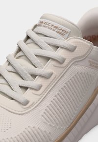 Ανοιχτό μπεζ αθλητικό παπούτσι Skechers BOBS με υφασμάτινη πλέξη υφής, μαξιλαρωμένη γλώσσα και λευκά κορδόνια δεμένα από πάνω.