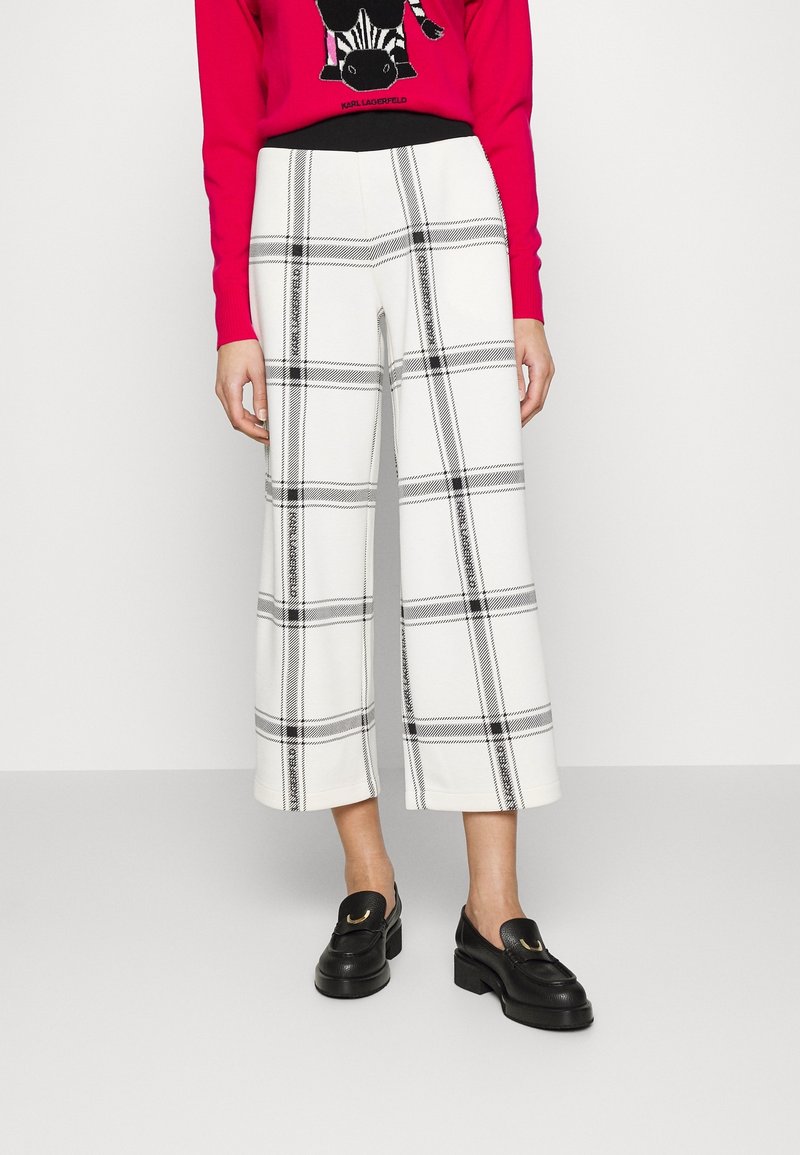 Białe spodnie o szerokich nogawkach w czarną kratę, z wysokim stanem i skróconą długością, zestawione z czarnymi mokasynami.