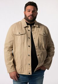 Béžová denimová bunda s knoflíkovým zapínáním vpředu, dvěma náprsními kapsami a volným střihem. Nositá přes černé tričko, se stříbrnými kovovými detaily.