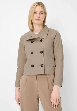 Woman wearing a beige cropped double-breasted jacket with black buttons and matching beige high-waisted trousers against a white background.