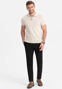 Beige Poloshirt mit kurzen Ärmeln und strukturierter Strickware, mit einer Drei-Knopf-Leiste, kombiniert mit schwarzen Hosen und braunen Slippern.