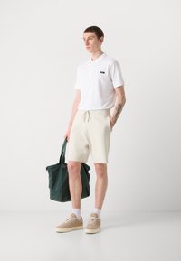 Camiseta polo blanca con un pequeño logo, pantalones cortos de punto beige y zapatillas color arena. Sosteniendo una bolsa de tote de color verde oscuro, de pie sobre un fondo sencillo.
