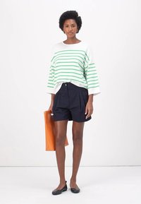 Pull oversized rayé blanc et vert, short plissé marine, ballerines noires et un tote bag orange sur un fond neutre.