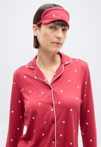Femme portant un pyjama rouge à motifs de cœurs et un masque de sommeil rouge assorti sur le front, sur un fond clair uni.