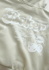 Olivovo zelená mikina s bielym grafikou. Dizajn obsahuje čerešne, mašľu a trblietky. Obsahuje nápis značky "Baker" prominentne zobrazený.