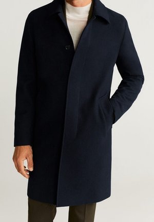 Mies pukeutuneena tummansiniseen yksiriviseen nappitakkiin, jossa on kaulus, valkoisen pooloneuleen päällä, yksi käsi taskussa, seisomassa neutraalia taustaa vasten.