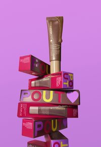 Tube de traitement des lèvres avec une finition dorée, empilé sur des boîtes rouges vives étiquetées "POUT," avec des accents multicolores, sur fond rose.