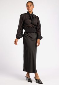 Zwarte doorschijnende blouse met een striknek, gecombineerd met een satijnen zwarte rok. Inclusief gerimpelde taille en puntige hakken met versieringen.