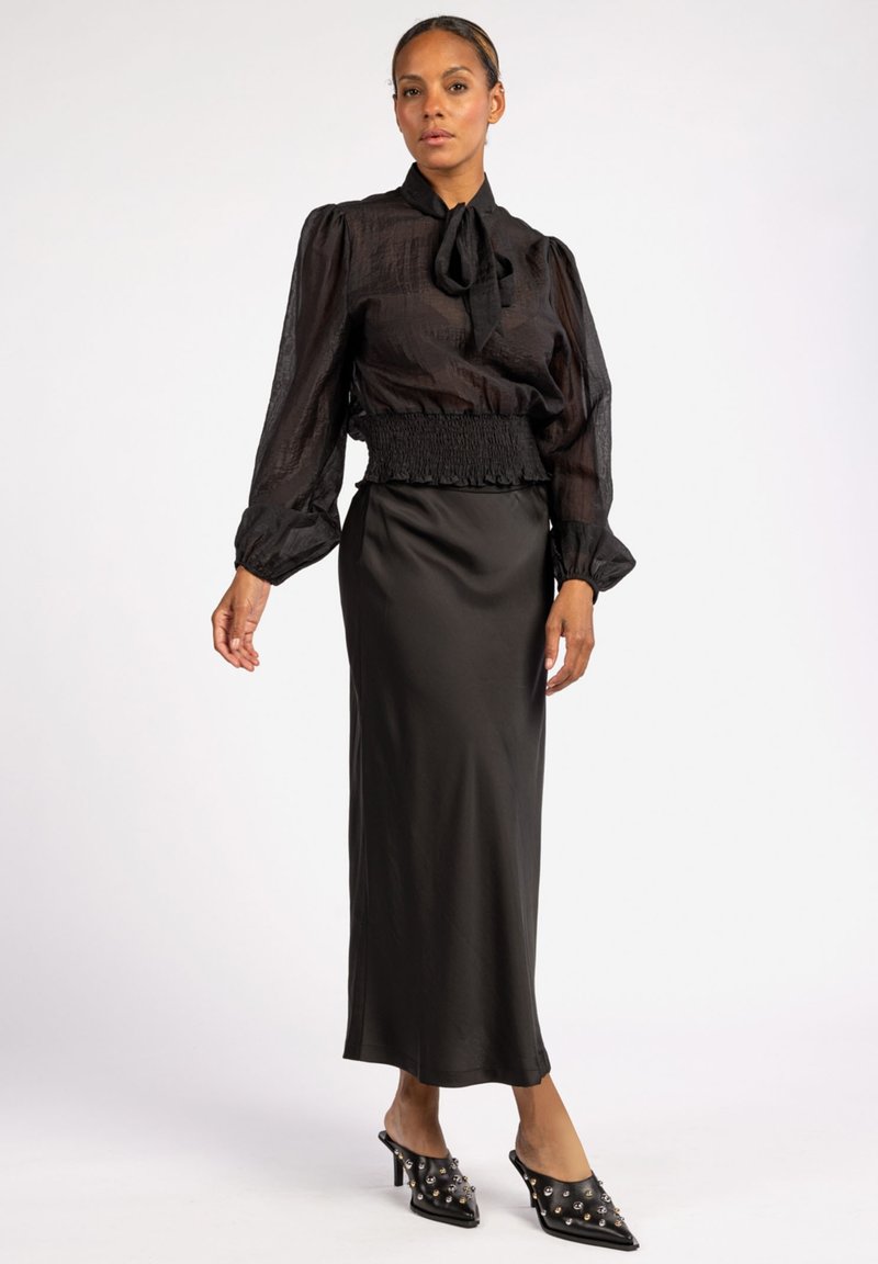 Zwarte doorschijnende blouse met een striknek, gecombineerd met een satijnen zwarte rok. Inclusief gerimpelde taille en puntige hakken met versieringen.