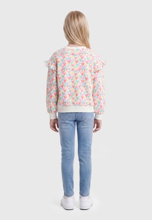 Dziecko z długimi blond włosami ubrane w pastelową bluzę w serduszka, jasnoniebieskie dżinsy oraz białe trampki, stojące tyłem.