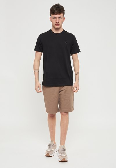 T-shirt noire à manches courtes avec logo sur la poitrine, associée à un short beige. Le modèle porte des baskets claires avec des accents gris et beige.