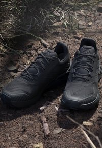 Chaussures de sport noires avec un dessus en maille texturée, une semelle robuste et un système de lacets bien visible, sur un fond de sol naturel.