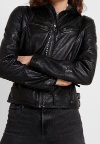 Veste en cuir noir avec fermeture éclair sur le devant, plusieurs poches zippées, détails matelassés et boucles latérales ajustables. Texture lisse.