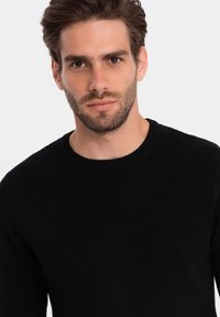 Schwarzer Strickpullover mit rundem Ausschnitt und strukturierter Optik. Nahaufnahme konzentriert sich auf Passform und haptische Details.