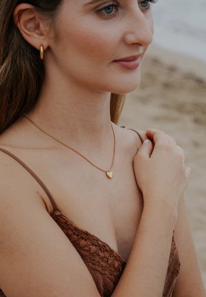 Náhrdelník s príveskom v tvare zlatého srdca na jemnom retiazke, nosený s hnedým čipkovaným topom, ktorý dopĺňa tón pokožky. Neformálne plážové prostredie.