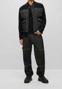 Giacca a vento nera con maniche in maglia, chiusura con zip e due tasche frontali, abbinata a pantaloni cargo in denim scuro con cuciture a contrasto e calzature nere.