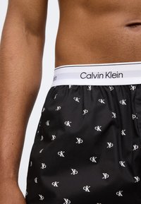 Μαύρα μπόξερ με λεία υφή που διαθέτουν διασκορπισμένα λευκά λογότυπα "CK". Η μέση είναι λευκή με τη λέξη "Calvin Klein" σε μαύρα γράμματα.