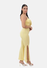 Jumpsuit amarillo con ribetes y cintura ceñida, con aberturas en los lados. Combinado con sandalias transparentes de tacón alto. Modelo mostrado en vista de perfil.