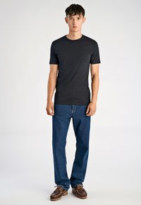 Maglietta nera a maniche corte in cotone, abbinata a jeans blu a gamba dritta e scarpe marroni con lacci, in piedi contro uno sfondo semplice.