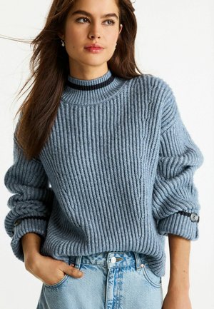 Hellblau gestrickter Pullover mit gerippter Textur, hohem Kragen und umgeschlagenen Bündchen mit schwarzen Akzenten. Kombiniert mit einer hoch taillierten Jeans.