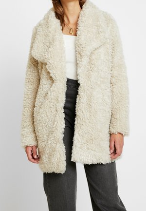 Mujer con un abrigo abierto de piel sintética esponjosa de color crema sobre una camiseta blanca acanalada y vaqueros gris oscuro, con collares dorados en capas.