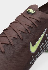 Artimas rudas tekstūruotas sportinis batelis su rudu batraiščiu, neoninės geltonos spalvos Nike Swoosh logotipu ir šešiakampiu "Kyrie" ženklinimu ant liežuvėlio.