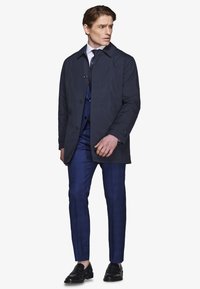 Mann trägt marineblauen Mantel über blauem Anzug mit weißem Hemd und Krawatte, schaut nach links, steht vor weißem Hintergrund.