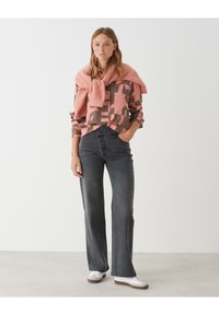 Camicia a fantasia mimetica in rosa e marrone, abbinata a jeans a gamba larga grigio scuro. Un maglione rosa pallido annodato attorno alle spalle. Sneakers bianche.