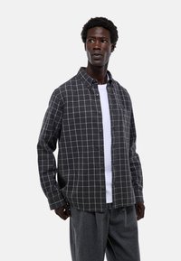 Chemise en flanelle gris à carreaux avec boutons, dotée d'un motif en grille blanche et d'une coupe décontractée. Portée par-dessus un t-shirt blanc uni.